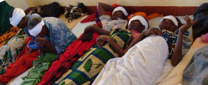 African women with bandages around their eyes lay on the ground after successful cataract surgery.